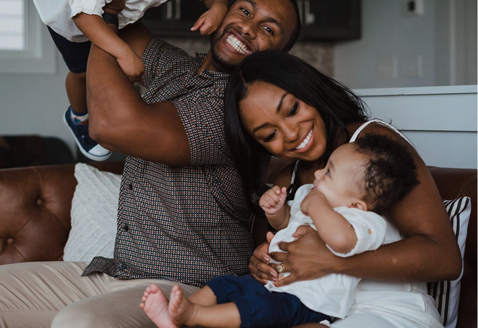 A father laughing while lifting his toddler overhead, next to a mother holding a baby, on a living room couch